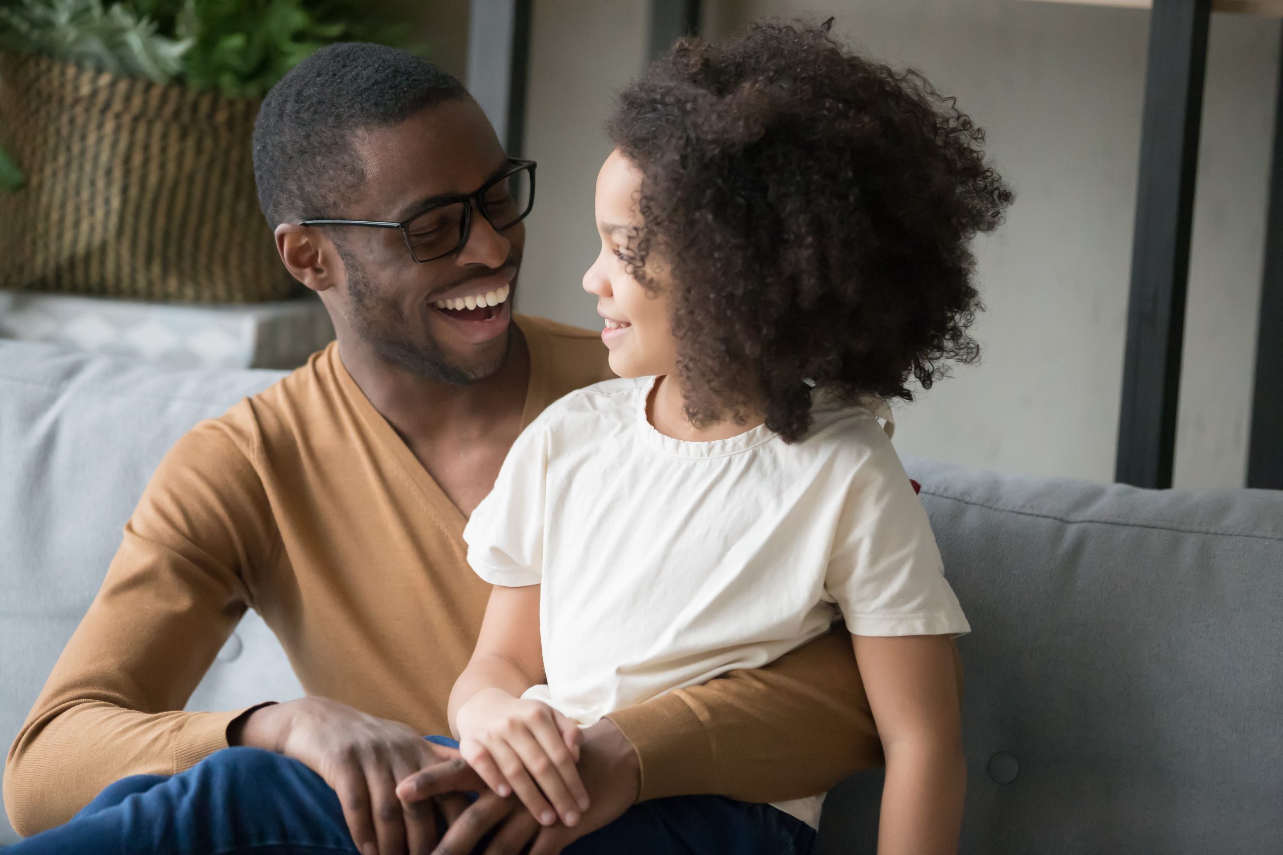 Smiling african American young dad sit on couch hug cute little daughter chatting or talking, happy black father spend time having conversation with funny preschooler child, enjoy weekend together